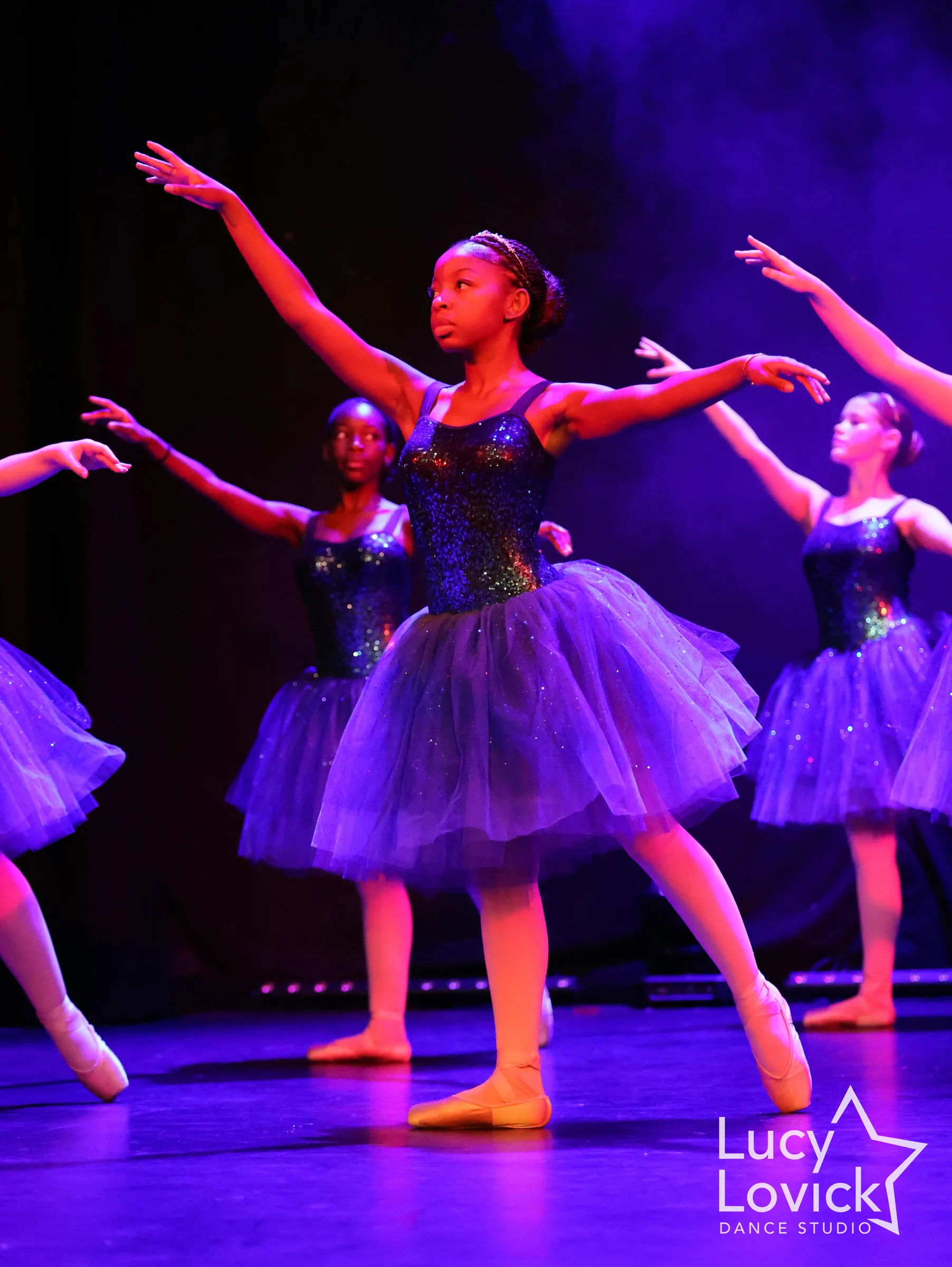 A young ballet dancer in a sparkling bodice and purple tulle skirt extends her arms with a focused gaze, other dancers softly blurred behind her.