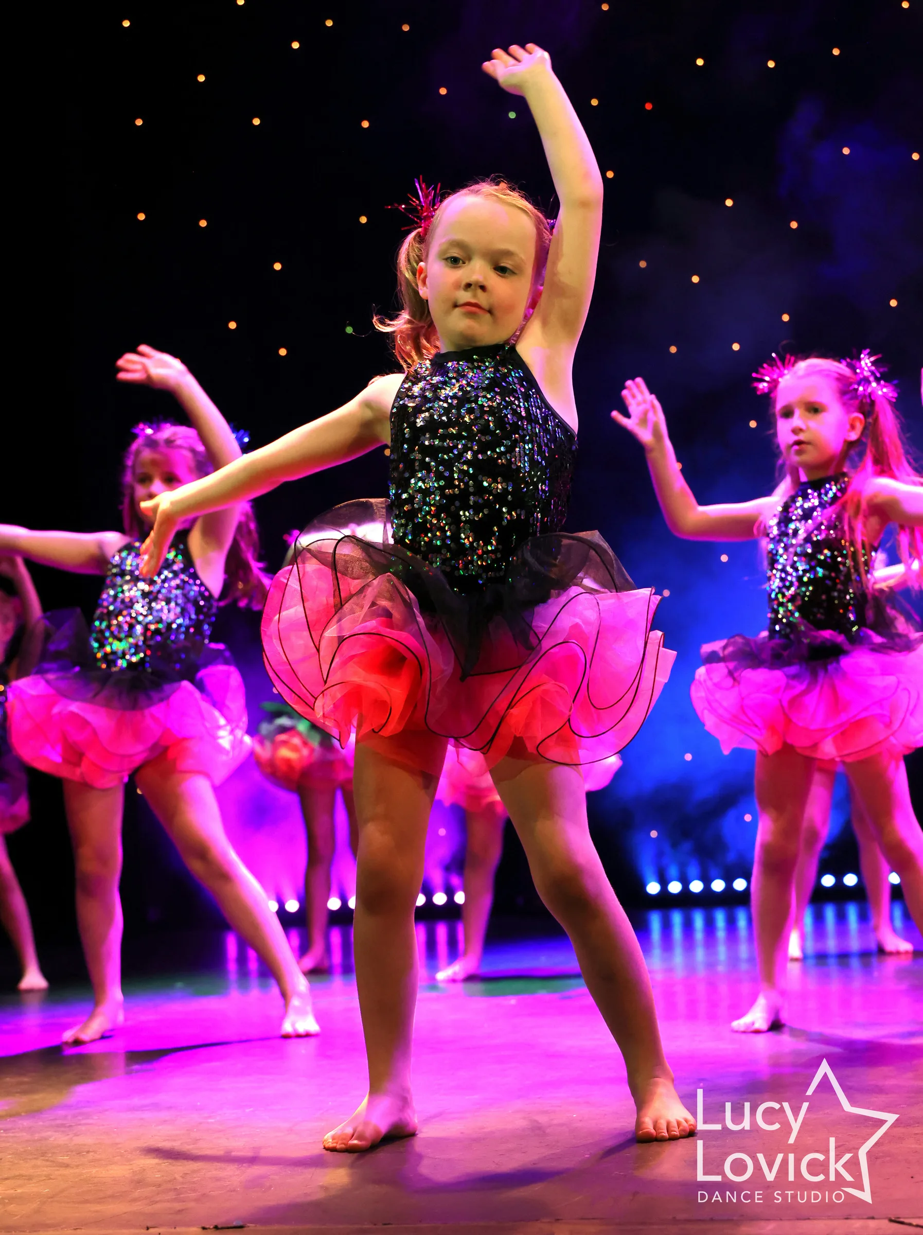 A little girl in a black sequinned top and bright pink tutu dances with one arm raised and a look of determined concentration.