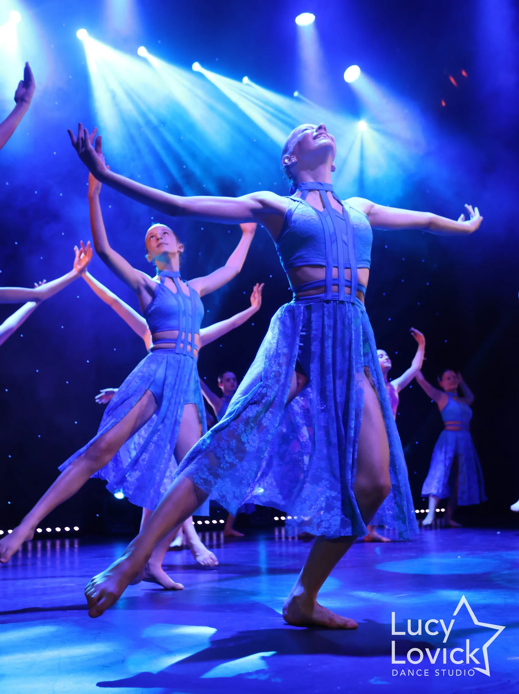 A dancer in a blue lace dress arches her chest towards the stage lights with arms spread wide, as other dancers reach upwards behind her in a blue haze.