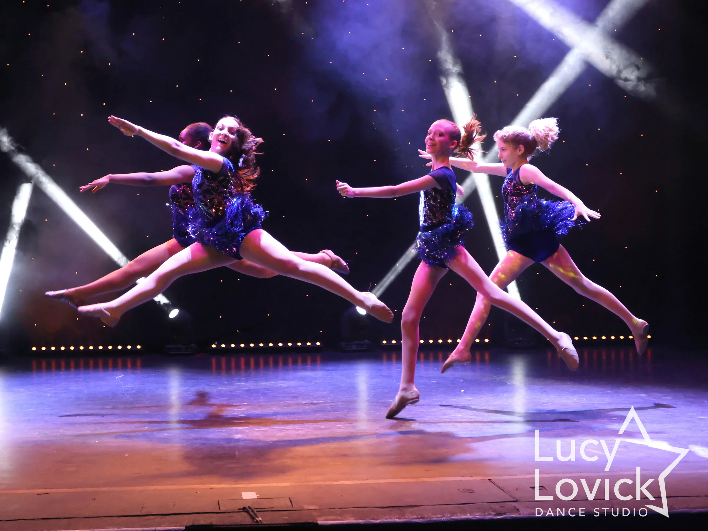 Young dancers in sparkly navy costumes soar through the air in joyful leaps, the girl in front grinning, as white beams cross behind them.