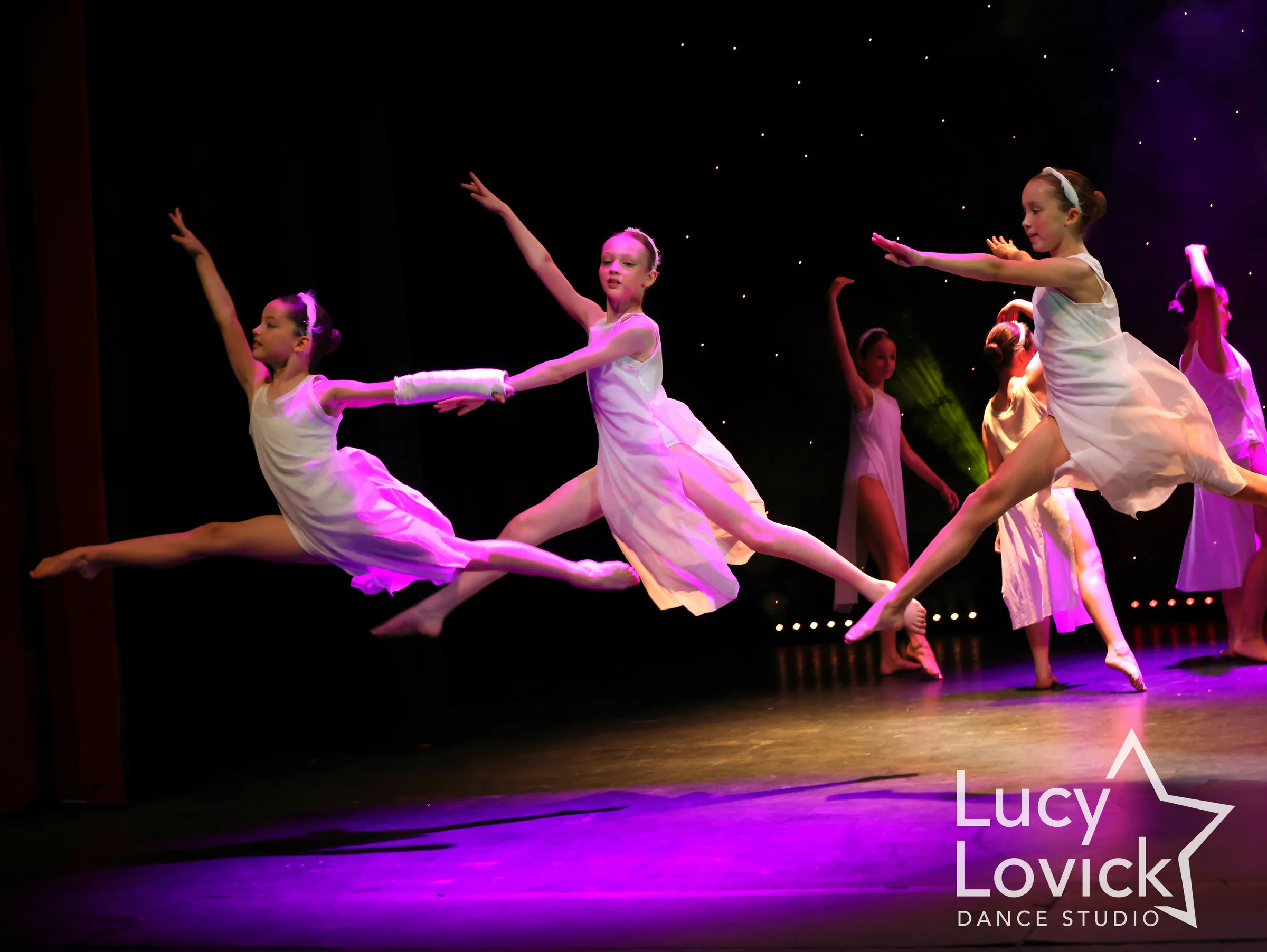 Young dancers in white flowing dresses leap through the air with legs outstretched, lit in magenta against a dark stage.