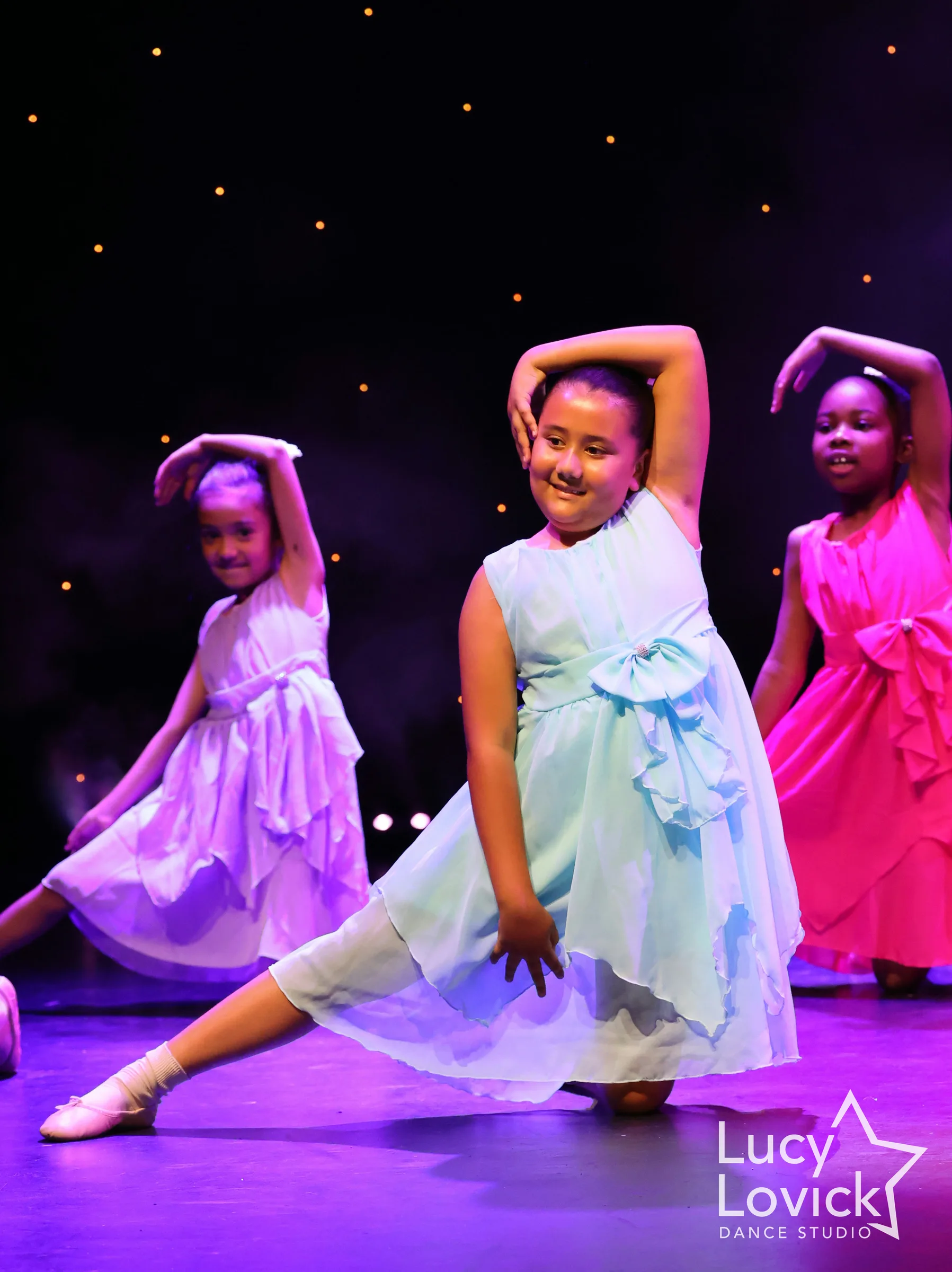 A young girl in a mint green party dress smiles as she lunges with one arm curved over her head, with girls in lilac and pink dresses behind.