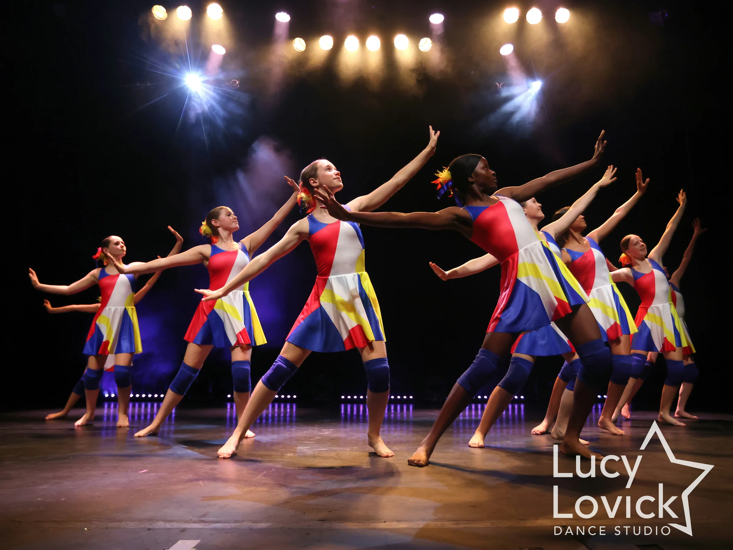 Teenage dancers in bright red, yellow and blue patchwork dresses reach their arms upward in a sweeping diagonal line beneath a rig of glowing stage lights.