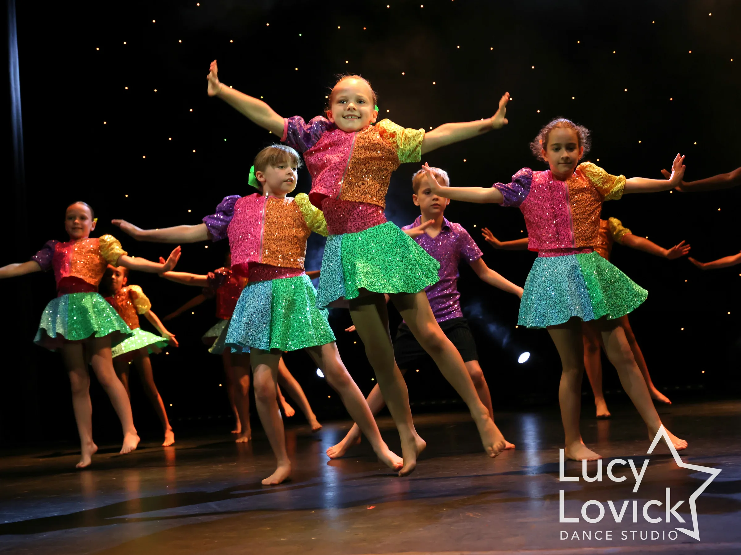 A beaming young girl in a rainbow sequinned top and sparkly green skirt dances with arms flung wide, surrounded by smiling classmates in matching costumes.