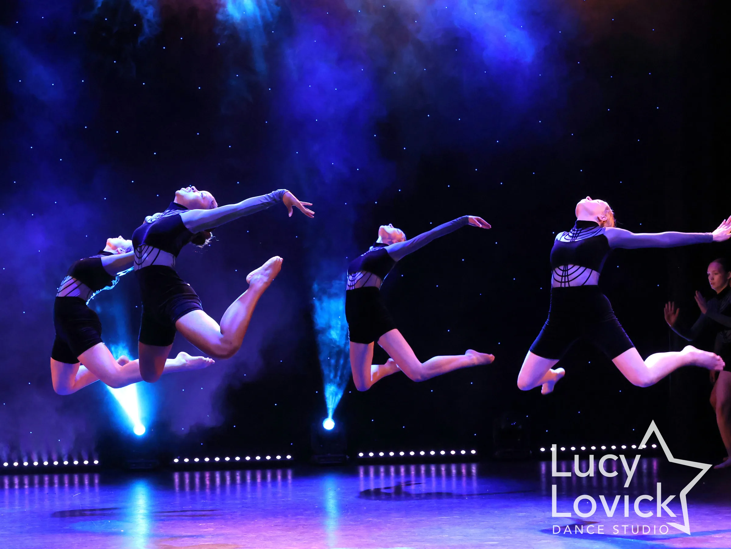 Four dancers in navy unitards soar through the air with heads thrown back and arms reaching forward, silhouetted against blue haze and a starry backdrop.