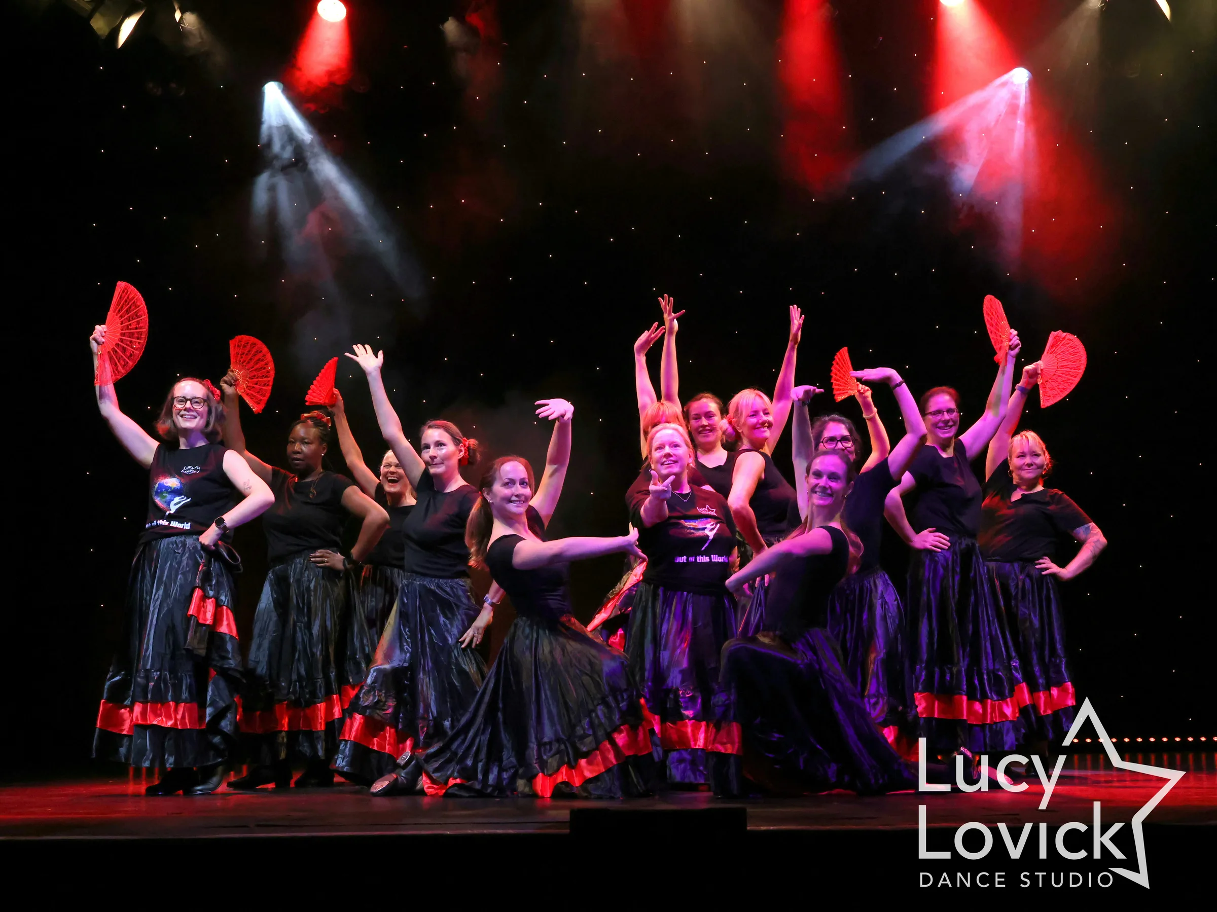 A smiling group of adult dancers in black tops and satin skirts with red trim hold up red fans in a celebratory finishing pose.