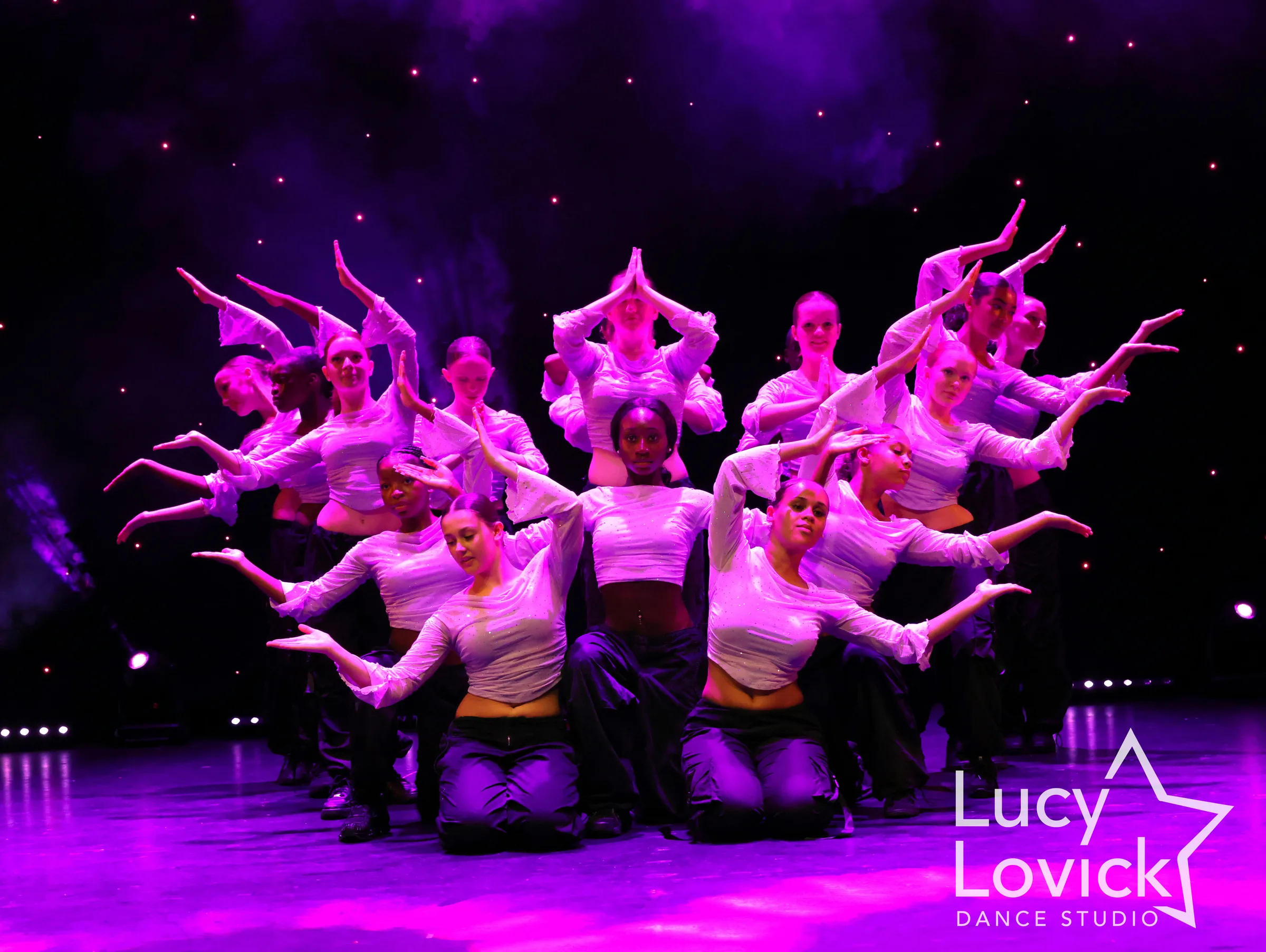 Teenage dancers in white crop tops and black trousers stack together in a layered formation, their arms fanning out in angular shapes under magenta light.